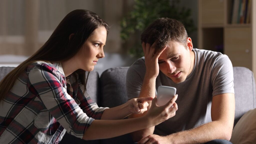 Admitted An Affair: man and woman sitting looking upset at the woman’s phone man looks guilty.