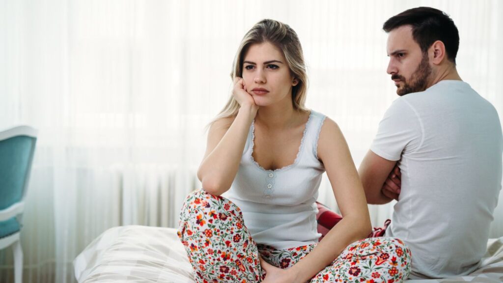 break up. Woman looking away sitting on the bed while the man sits behind her looking at her. They both look upset and angry