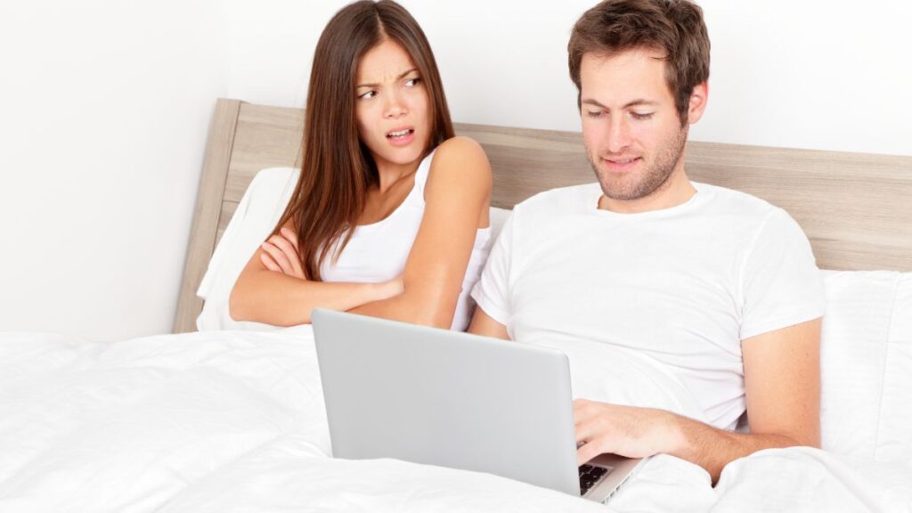 Cheater man smiling at his computer next to an upset woman in bed.