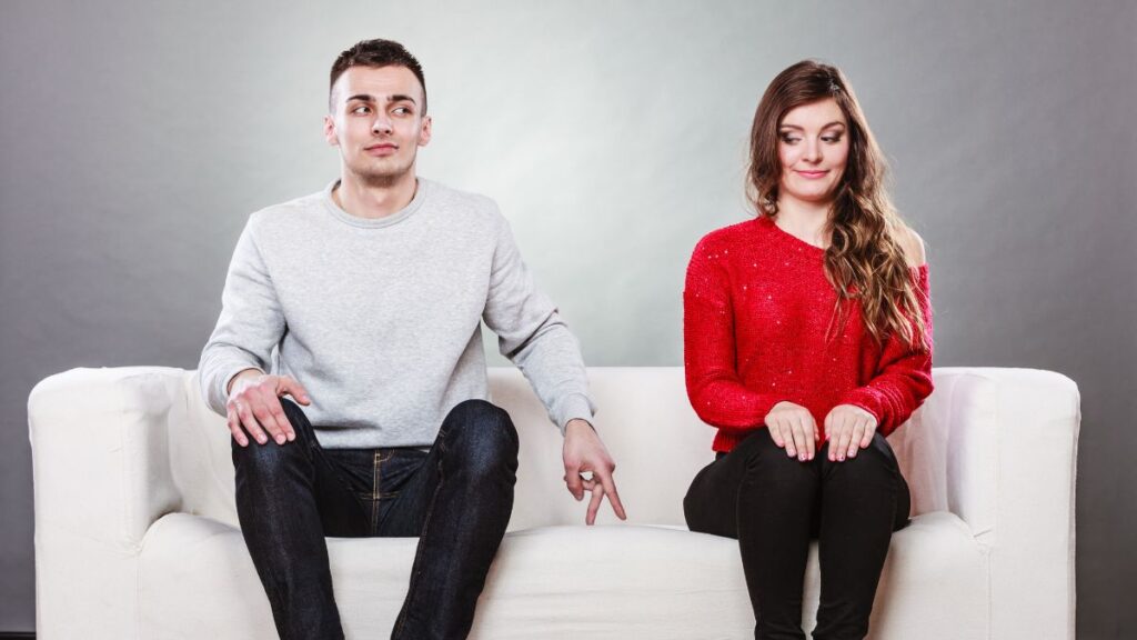 couple sitting on the couch. man is inching his fingers towards her and she's looking away -