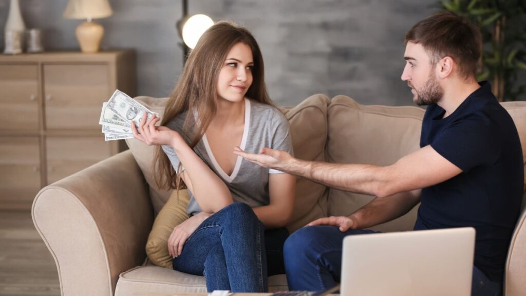 woman holding money while sitting on the couch keeping it away from boyfriend who is also on the couch