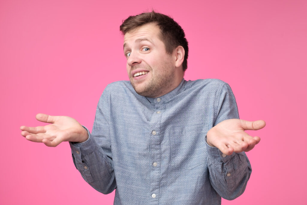 Uncertain young stylish man wears blue shirt, shrugs shoulders being puzzled or confused. Caucasian unsure male make gestures doubtfully with hands standing on pink background.