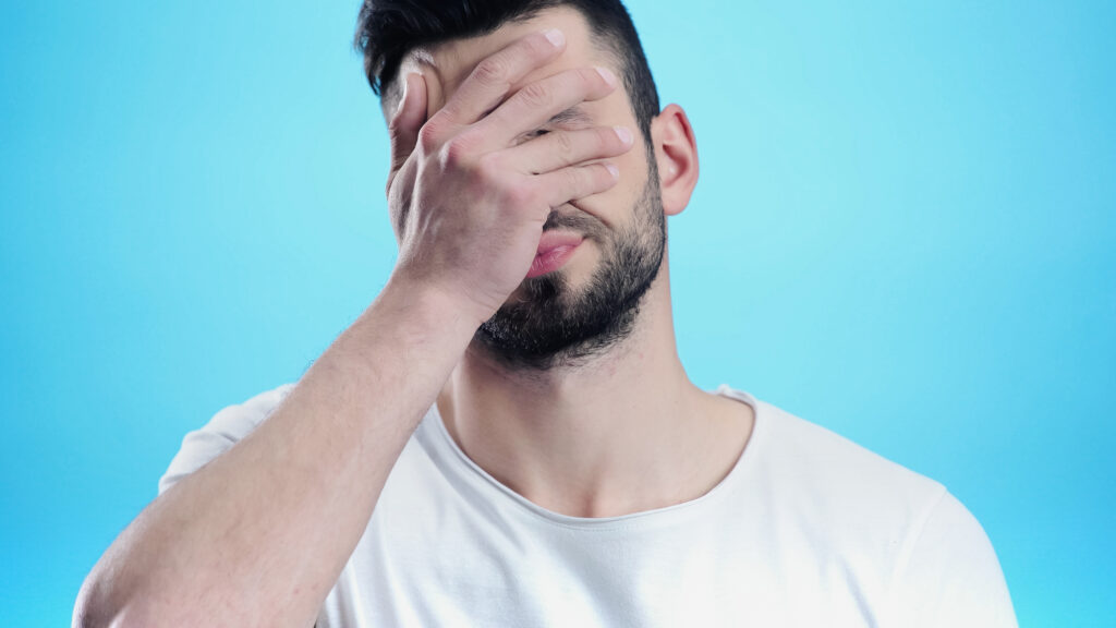 blue background, white man holding his face in disappointment. covering his eyes