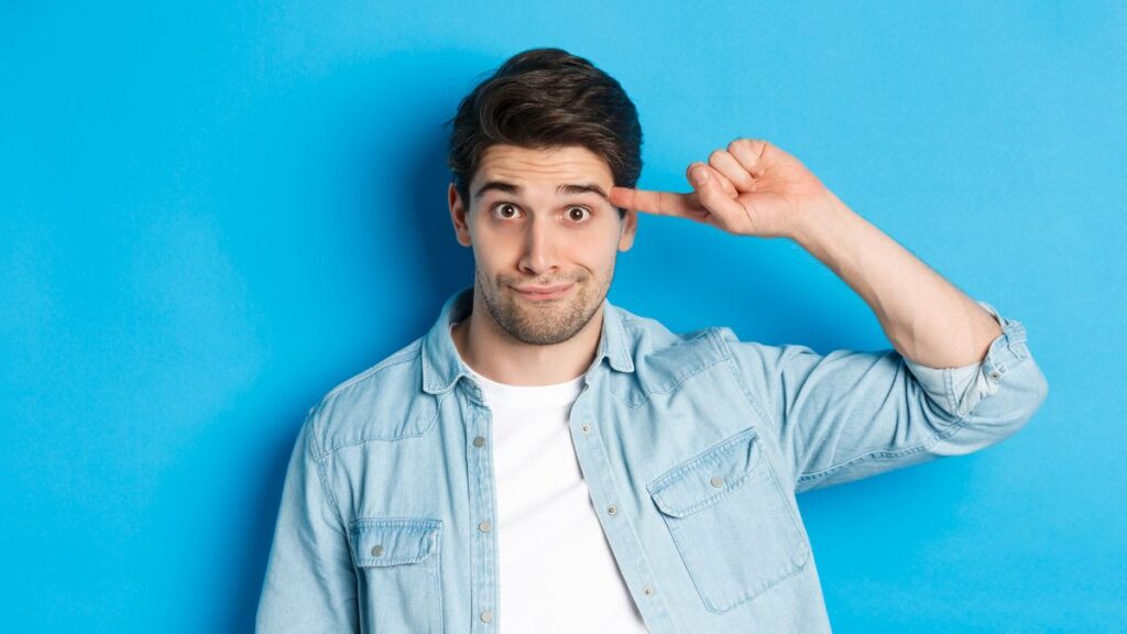 Dumb man standing and pointing to his head while making a goofy face.