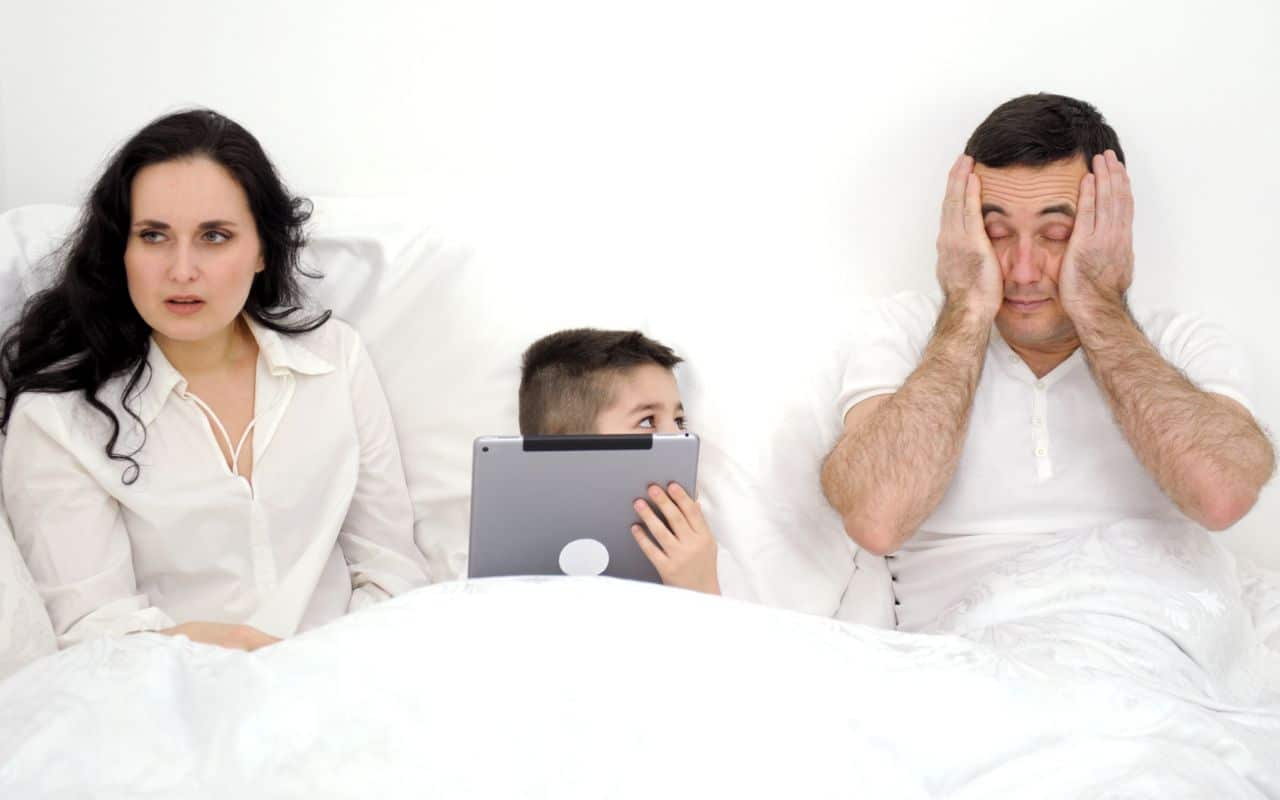 family in bed, child on ipad, parents look tired and miserable