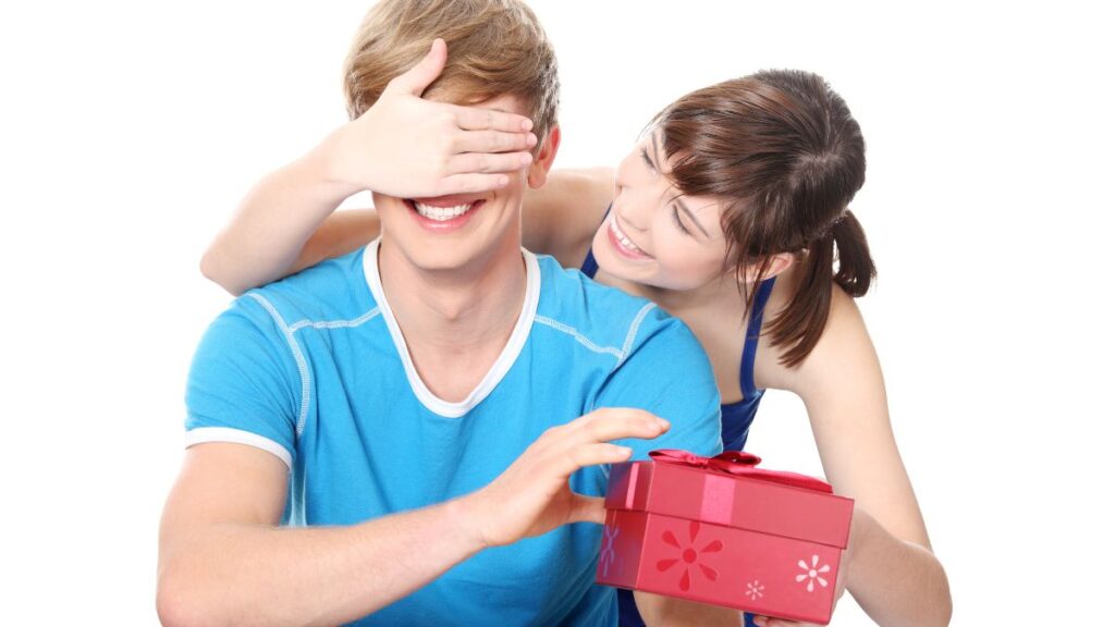 Gifts: woman covering mans eyes giving him a gift and leaning over his shoulders.