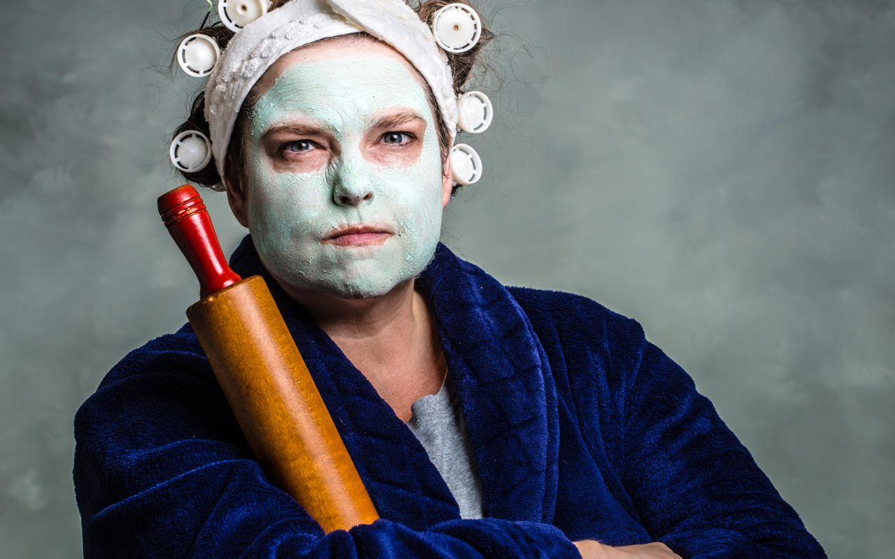 grumpy woman in a robe, face mask and hair rollers holding a rolling pin. 