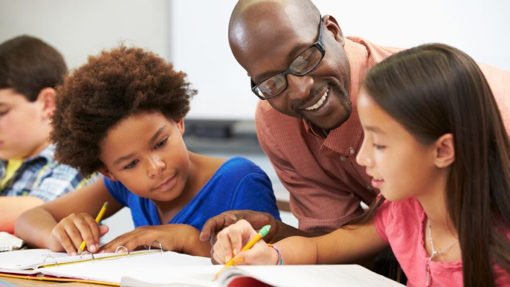 Male Elementary School Teachers: male teacher sitting with two students smiling and reading.