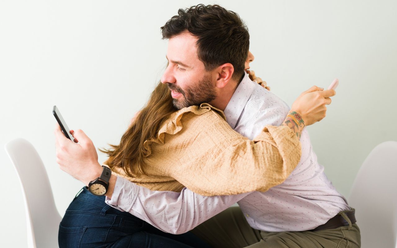 man and woman hugging and looking at their phones over the others' back