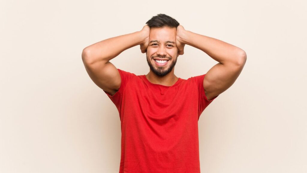 Man wearing a red shirt with his hands up to the side of his head smiling