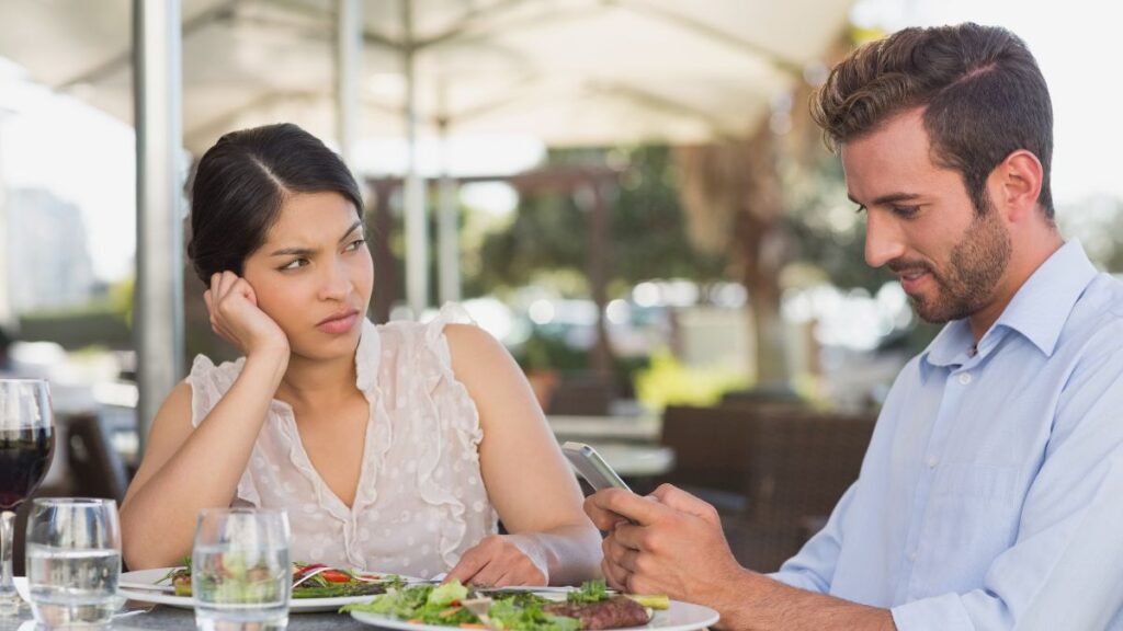 One-Sided man and woman sitting at a lunch table man on his phone woman looking at him upset.