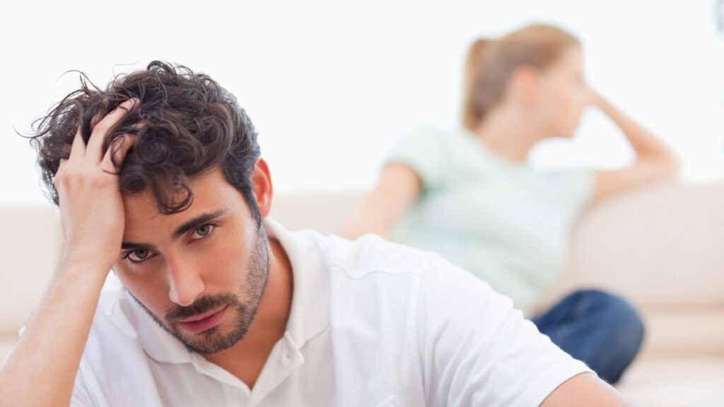 man looking upset with his hand on his head. Wife is behind him upse