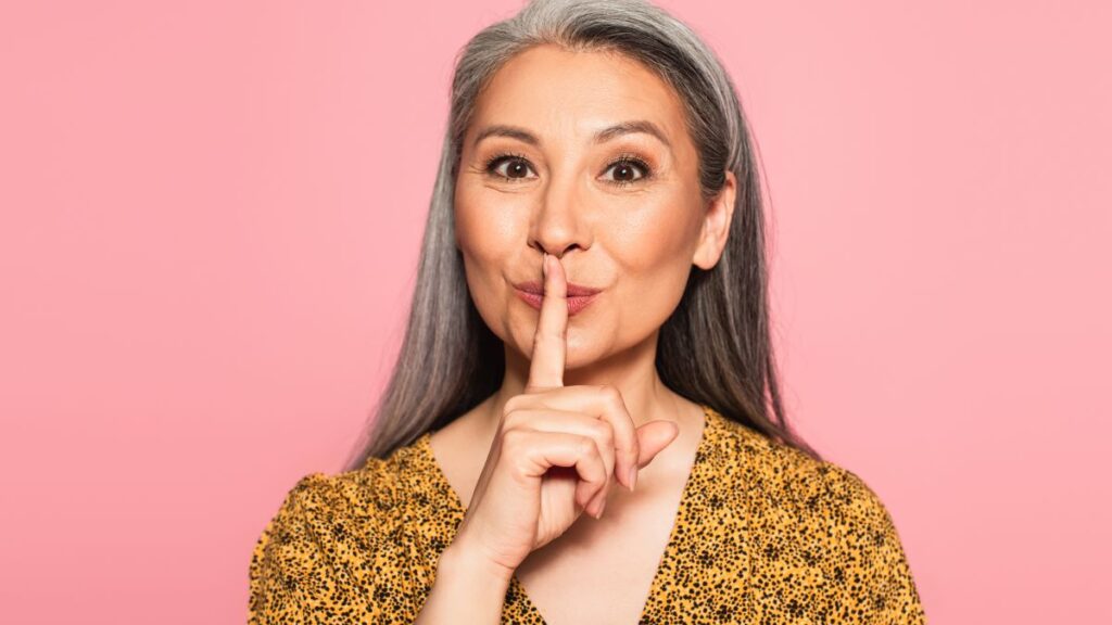 Softer Vice: woman with gray hair with her finger up to her mouth to signal quiet.