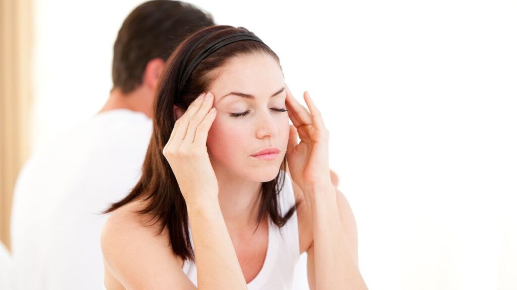 woman with eyes closed looking annoyed with hands on temples and man behind her